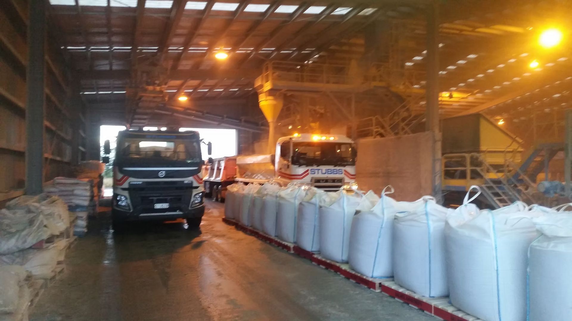 Truck inside a warehouse beside rows of white bulk bags under warm industrial lighting