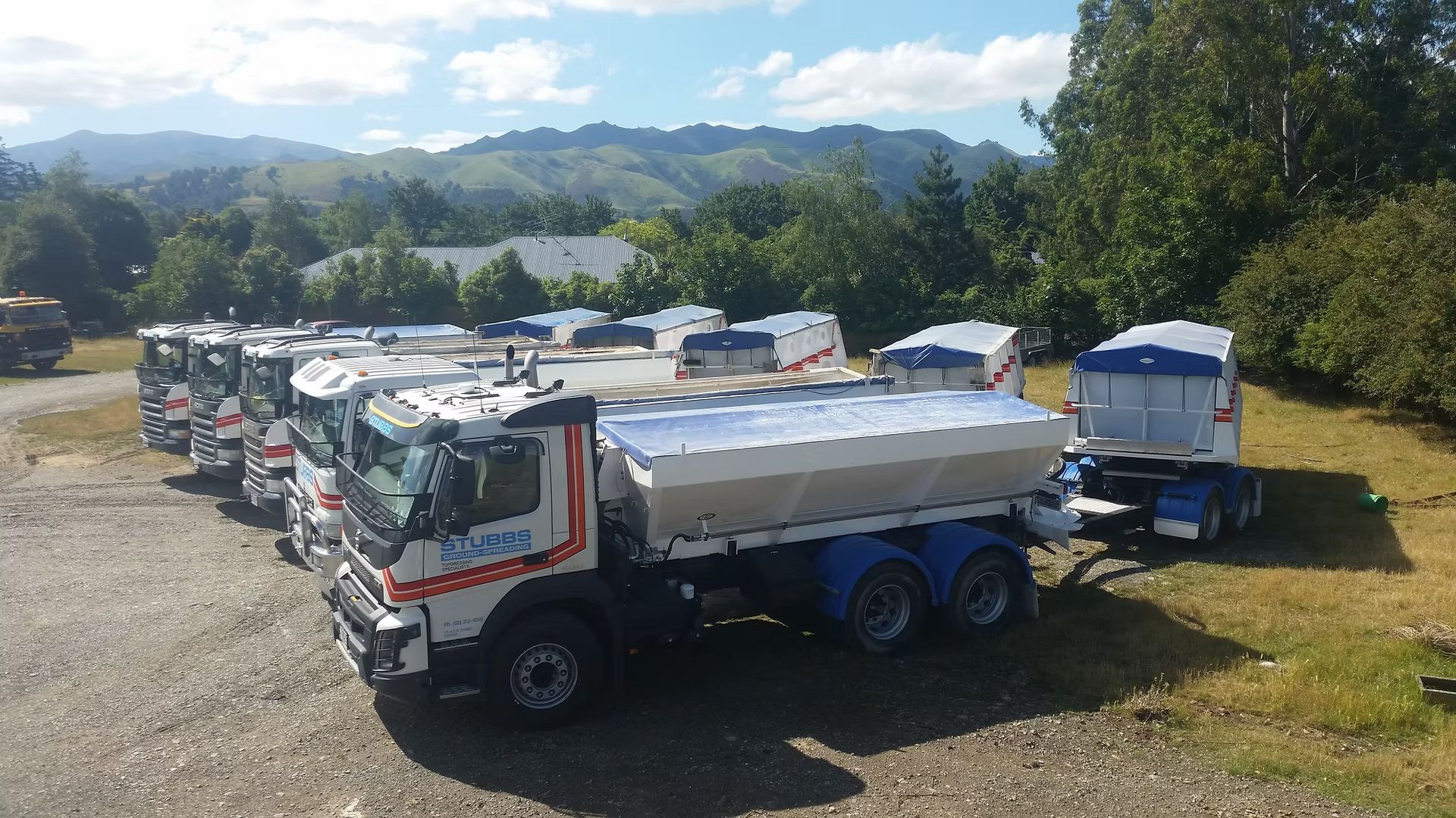 Line of large dump trucks parked on gravel in a sunny lot with hills and trees in the background