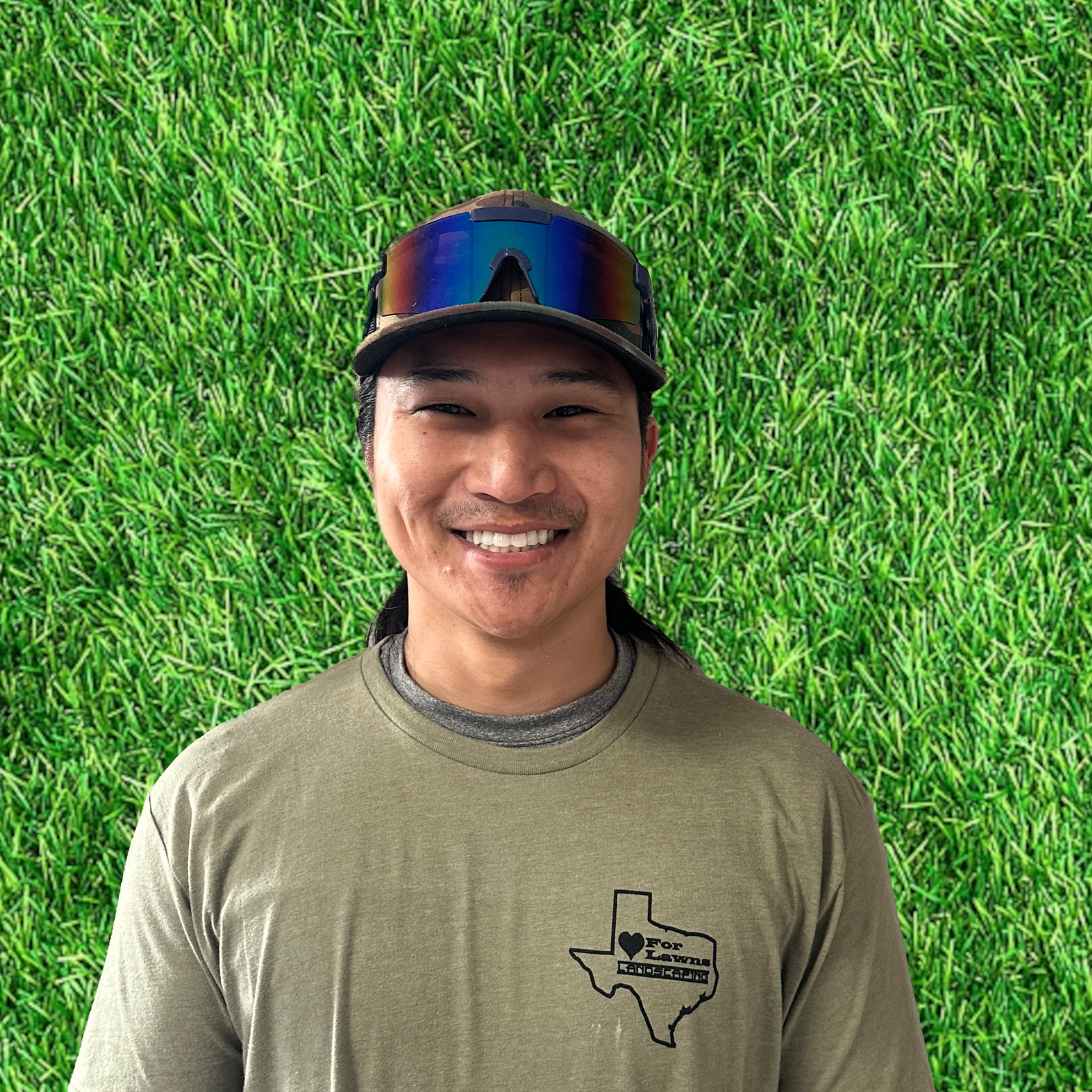 A man wearing a hat and sunglasses is smiling in front of a green field.