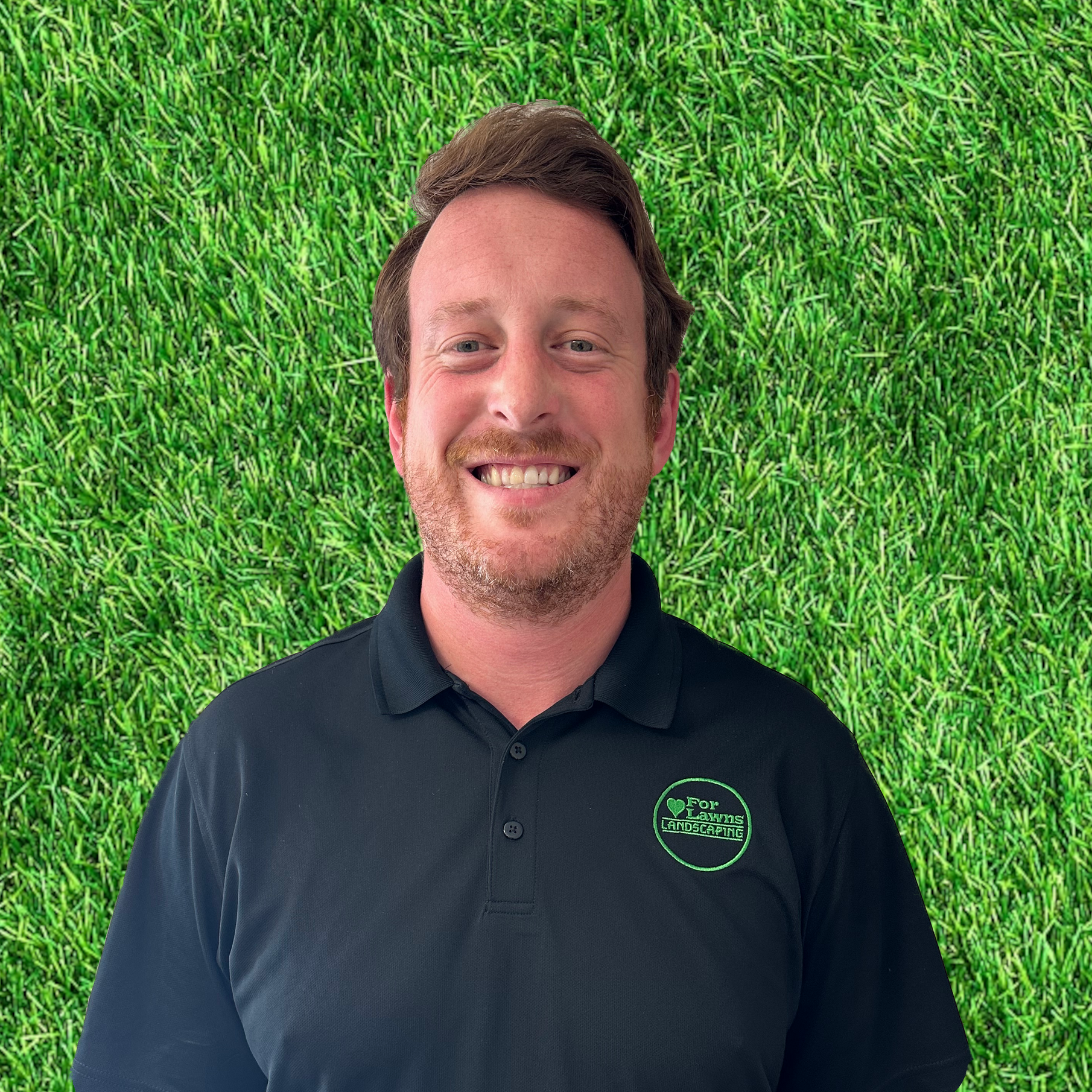 A man in a black polo shirt is smiling in front of a green grass background.