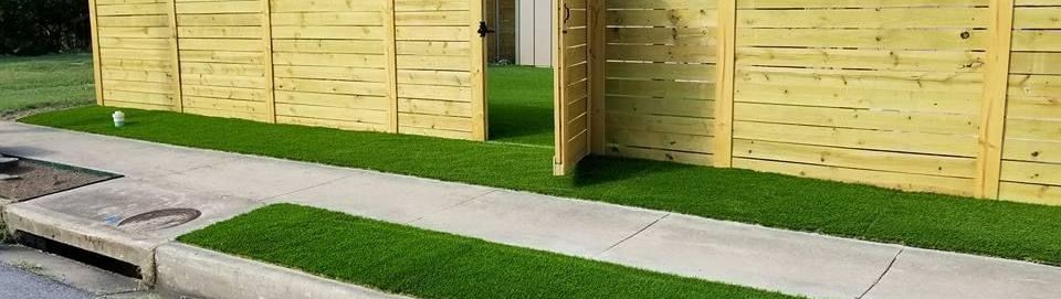A wooden fence is surrounded by artificial grass and a sidewalk.