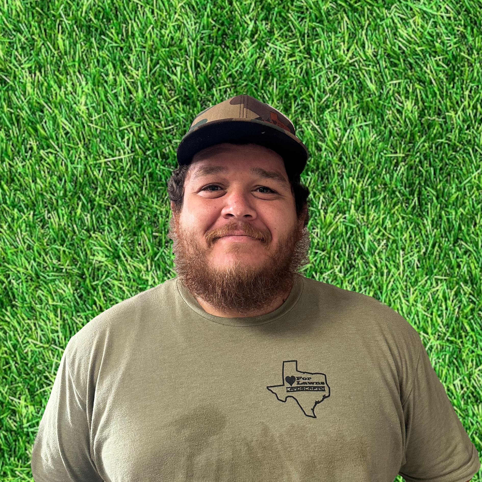 A man with a beard wearing a hat and a t-shirt is standing in front of a field of grass.