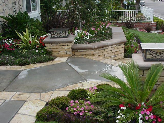 Stone patio garden with curved flower beds, pink and red flowers, and a black metal bench