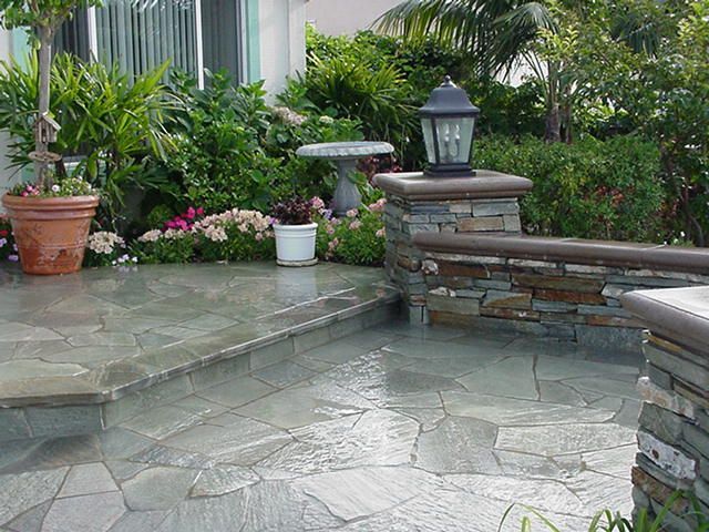 Stone patio with potted plants, lush greenery, and a lantern-topped retaining wall