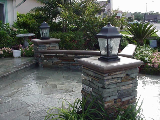 Stone patio with lantern-topped pillars, curved seating wall, and lush greenery in a landscaped garden.