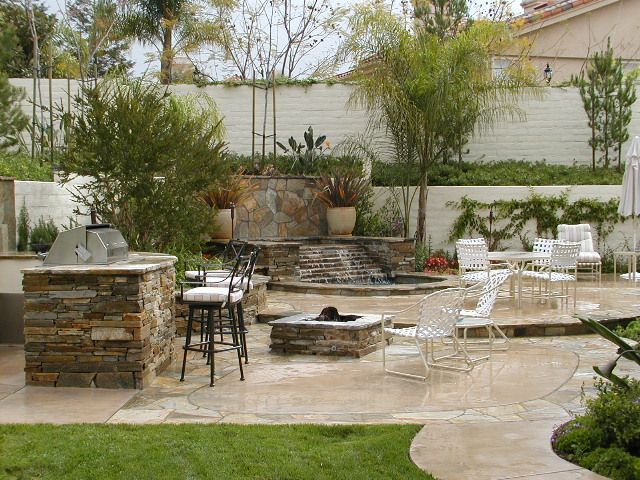 Stone patio backyard with outdoor seating, fire pit, and built-in stone grill surrounded by greenery