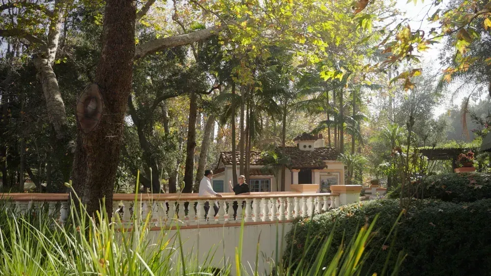 Tree-lined garden walkway with a white railing and a house in the background, dappled in sunlight