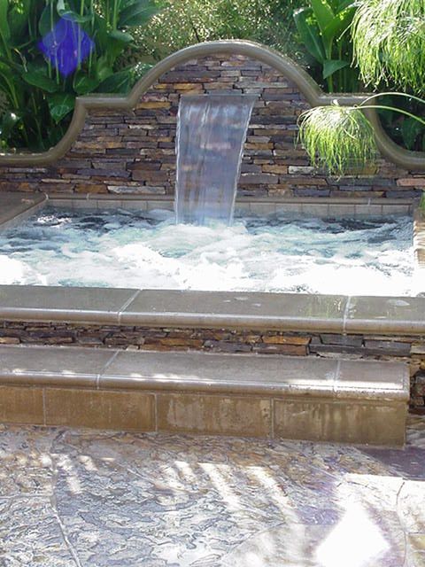 Stone fountain with water spilling into a shallow pool, surrounded by greenery and sunlit paving.
