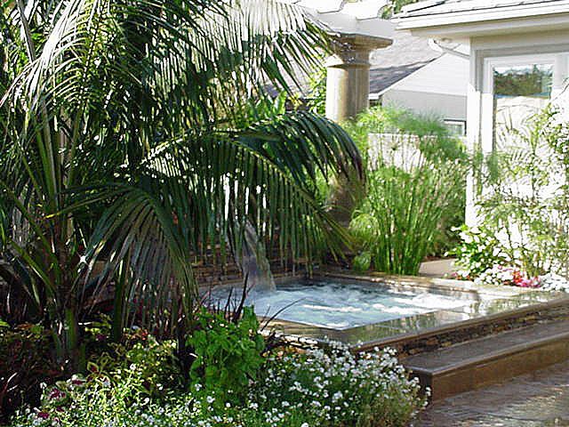 Tropical garden with palm trees, white flowers, and a small stone fountain beside a patio