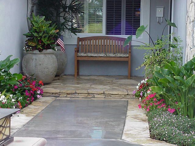 Front porch with wooden bench, large potted plant, stone walkway, and pink flowers by the entrance.