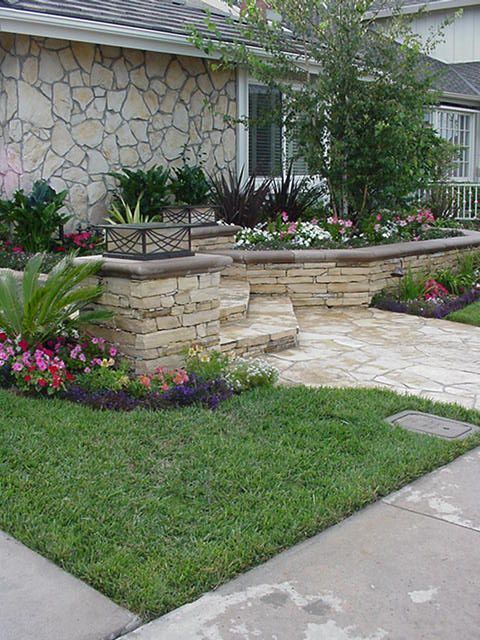 Landscaped front yard with stone retaining wall, flowers, shrubs, and a grassy lawn beside a house