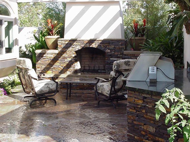 Outdoor patio with stone fireplace, cushioned chairs, and wet tiled floor in a landscaped backyard