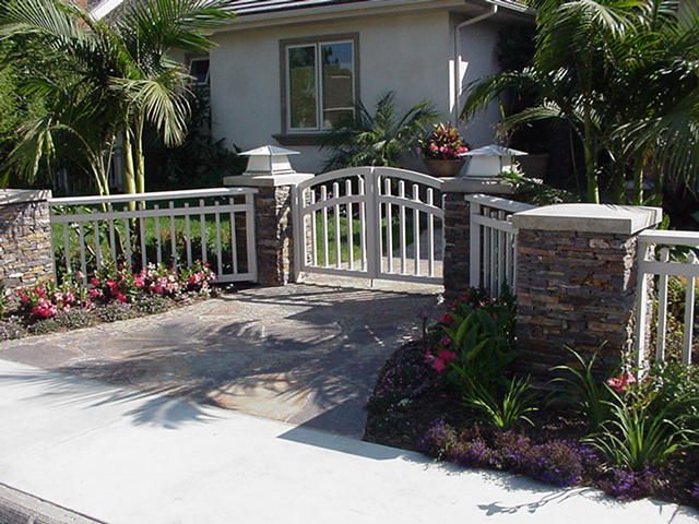 Front yard with a white gate, stone pillars, and tropical plants in front of a house