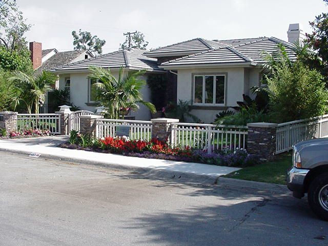 Single-story suburban house with white railing, red flowers, and a parked car on the street