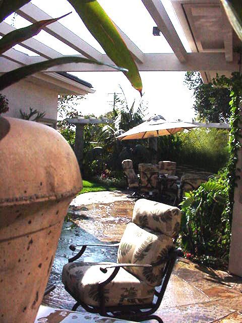 Patio with cushioned chairs, wet stone floor, potted plants, and an umbrella in a sunny garden