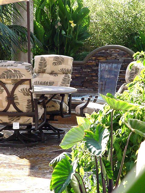 Patio seating with patterned chairs beside a small brick fountain, surrounded by lush green plants.