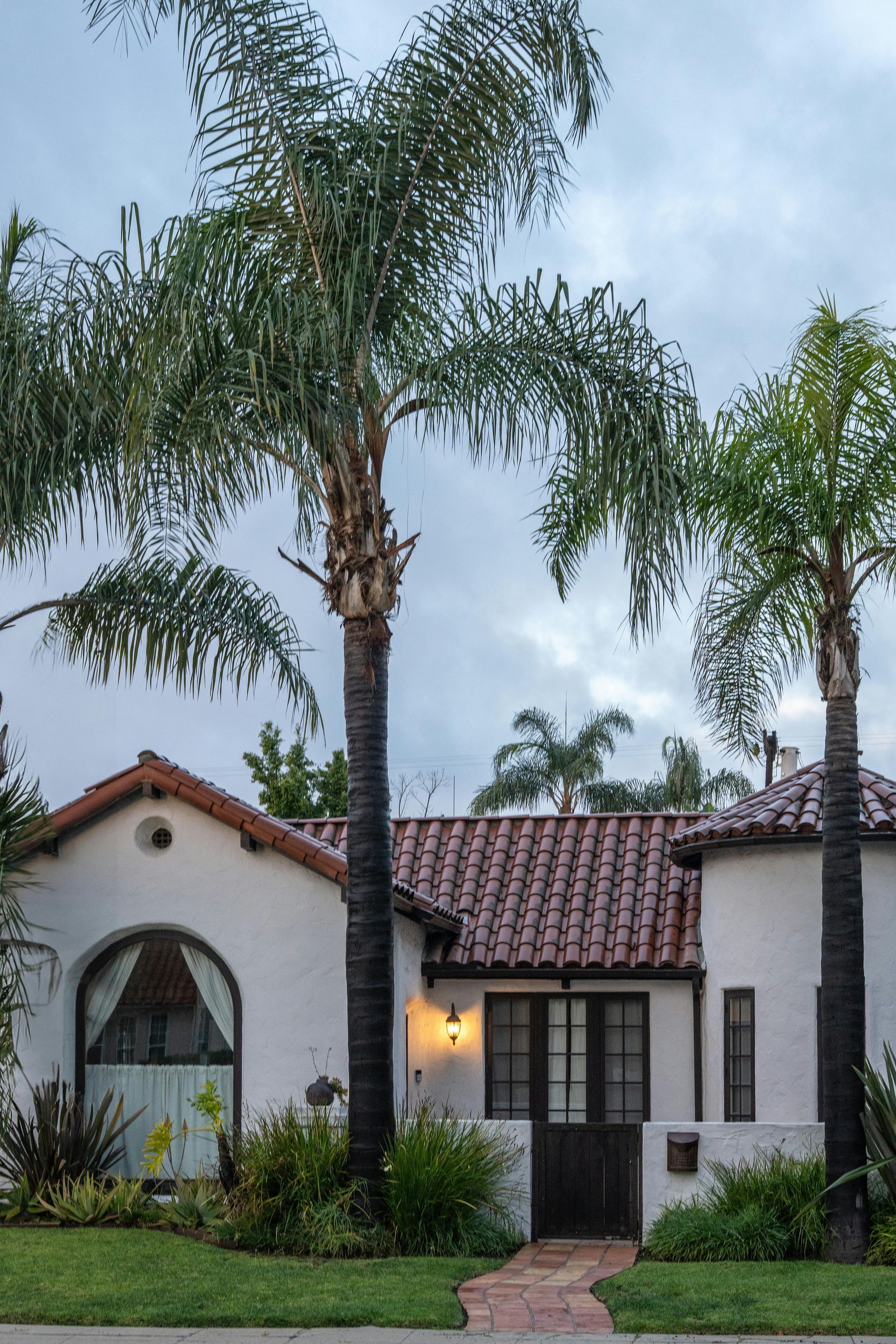 White stucco house with red tile roof and tall palm trees in front, lit at dusk
