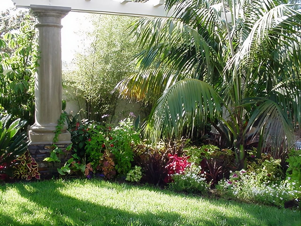 Lush garden with palm trees, flowering shrubs, and a stone column beside a green lawn