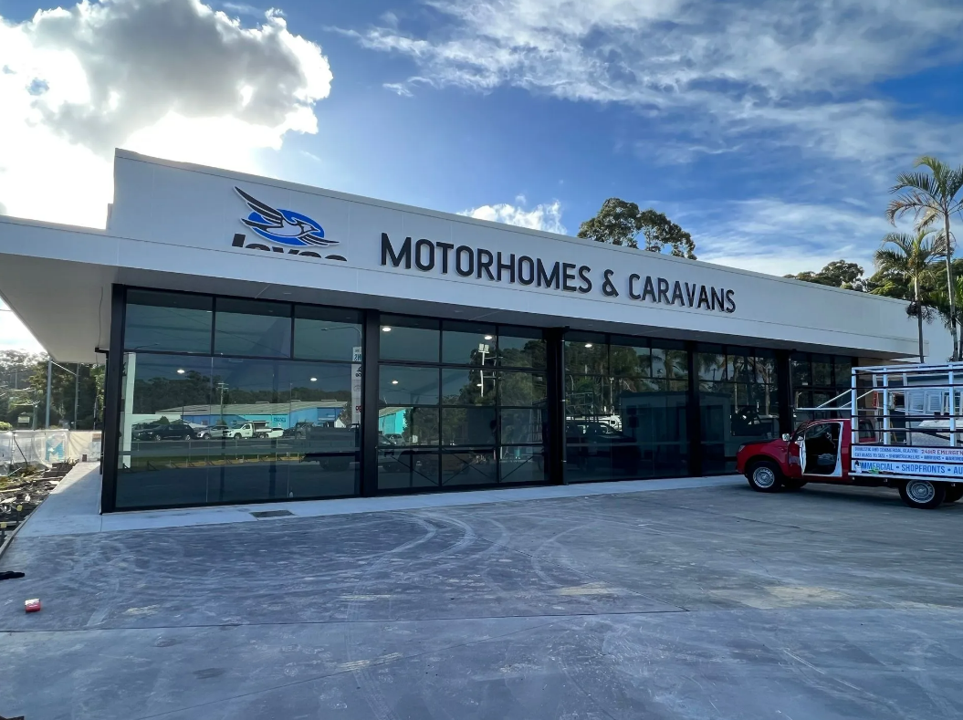 Motorhomes and caravans dealership with glass storefront under a blue sky; red truck parked outside
