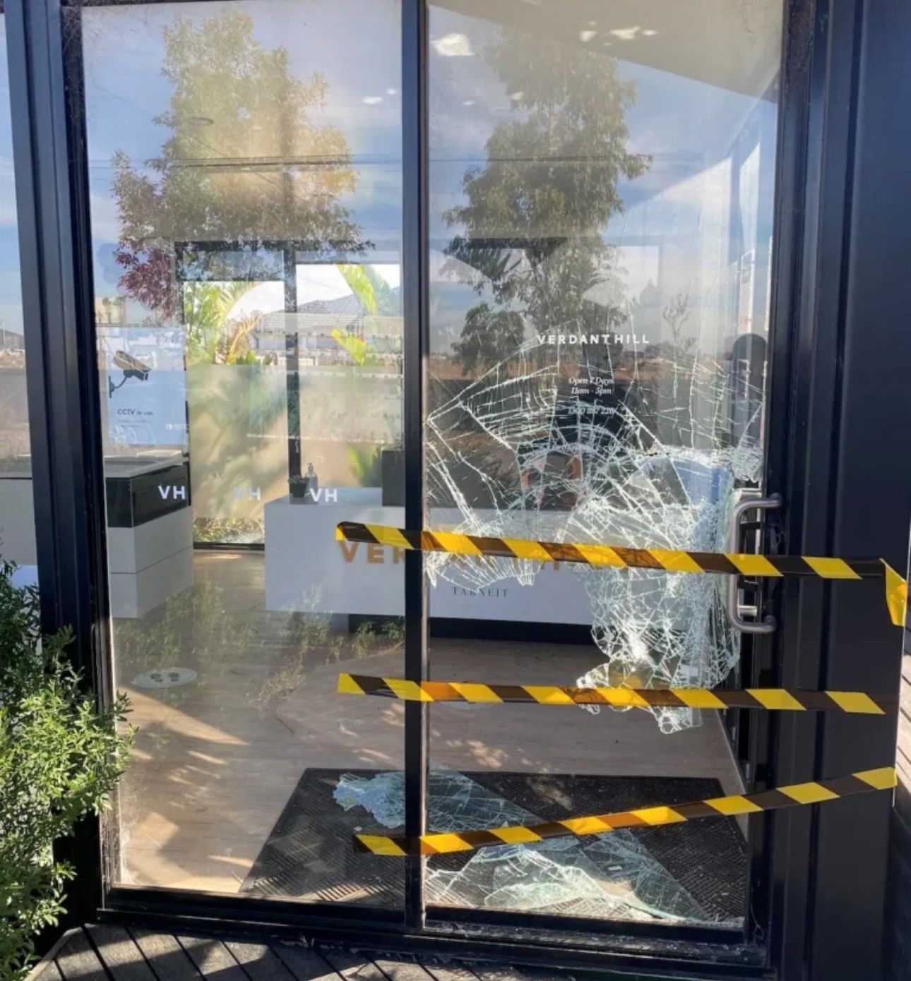 Storefront glass door with shattered pane, yellow caution tape, and reflections outside.