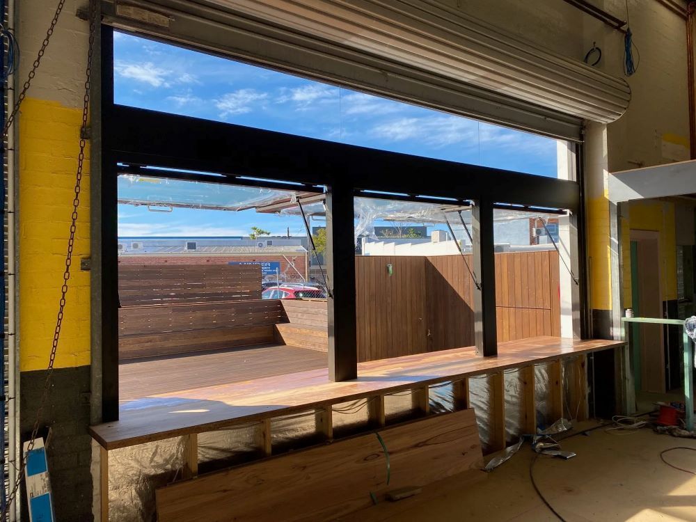 Storefront window with broken lower panels and construction debris inside, looking out to a fenced lot.