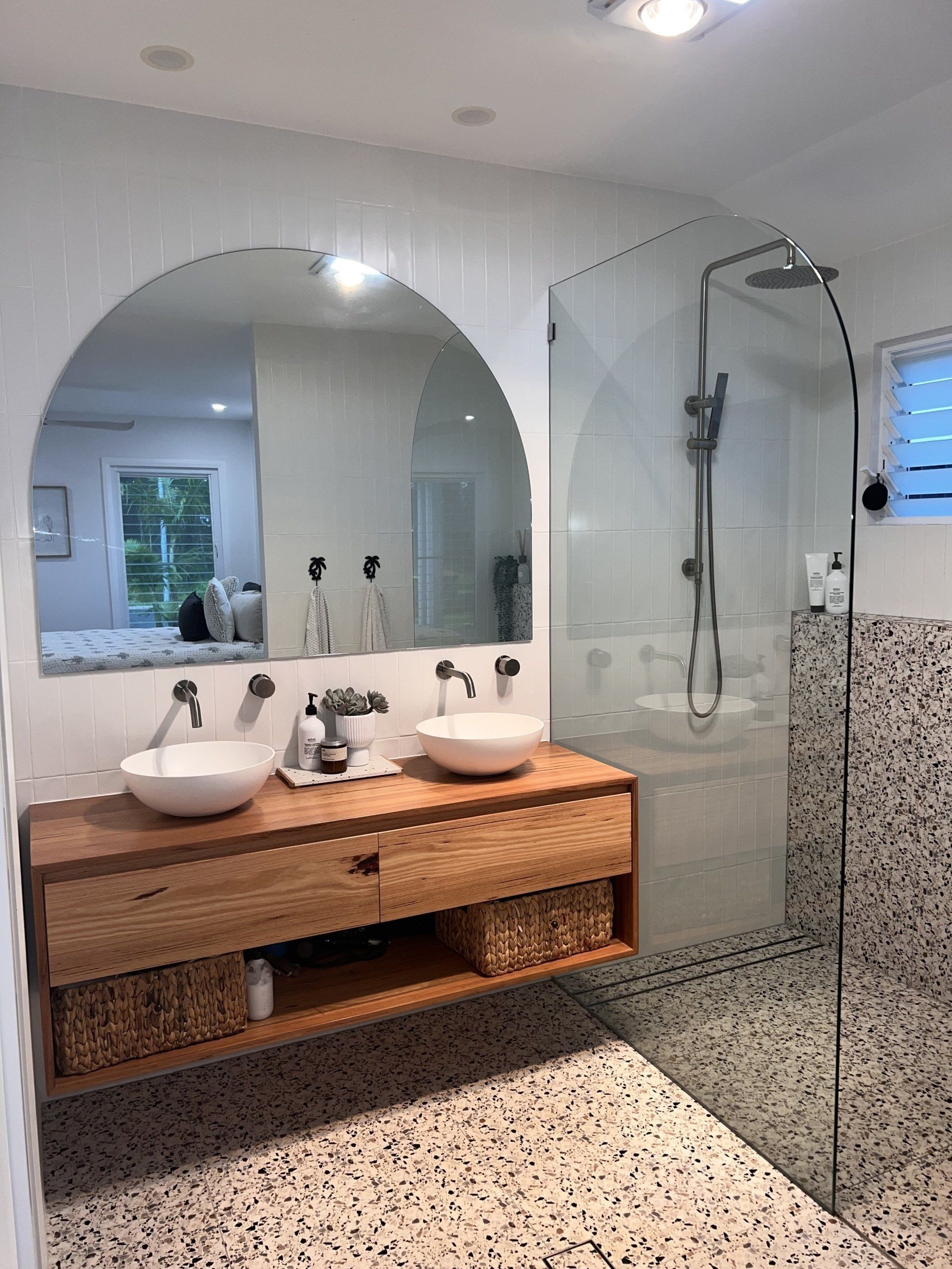 Modern bathroom with double sinks, arched mirror, glass shower, and speckled tile floor
