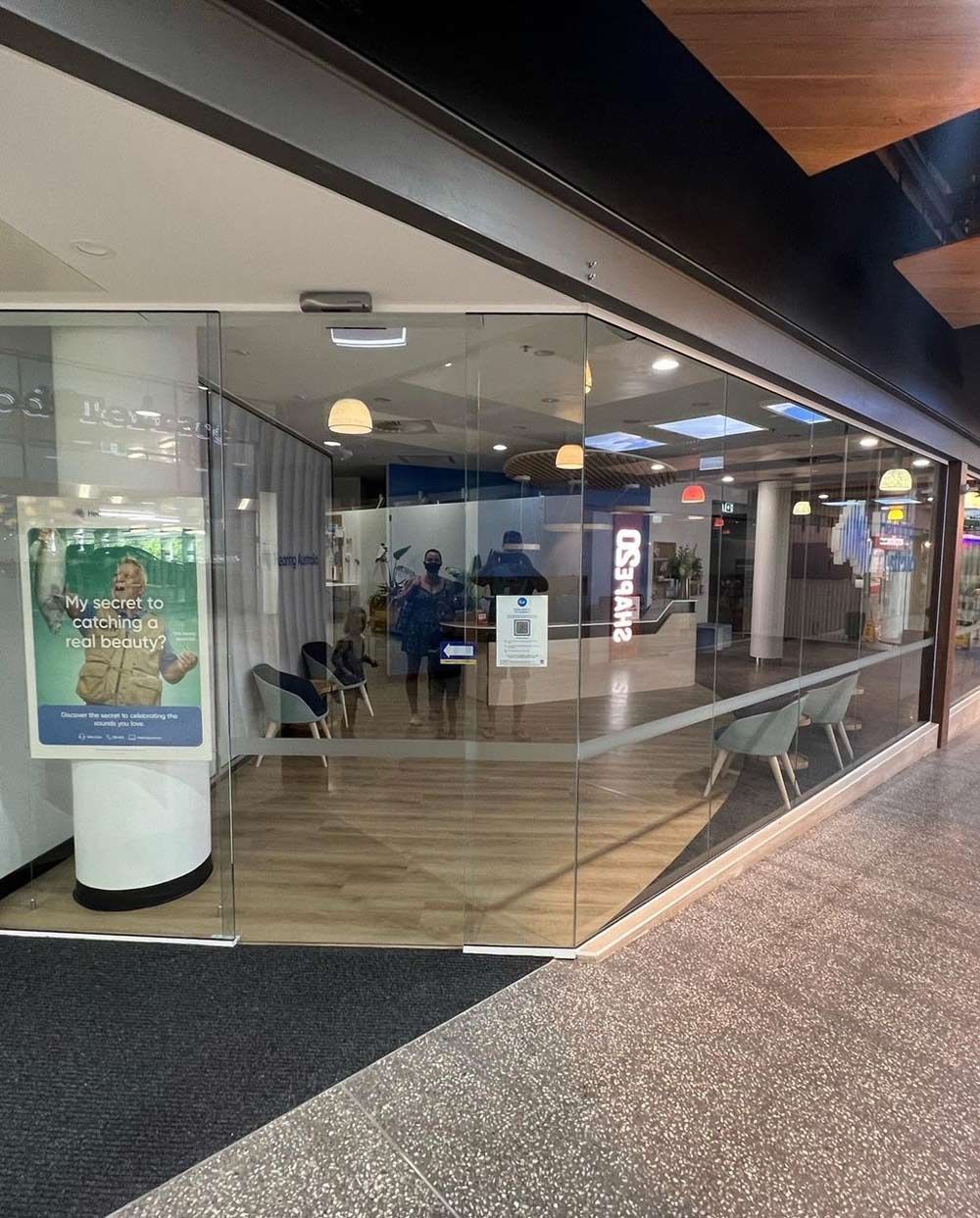 Empty mall corridor beside a storefront with glass windows, posters, and a blue-lit interior.