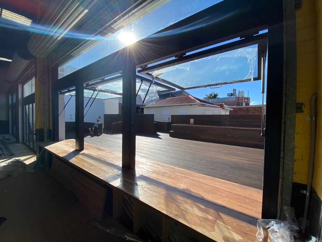 Covered outdoor patio with wooden deck, beams, and bright sunlight, overlooking blue sky and rooftops