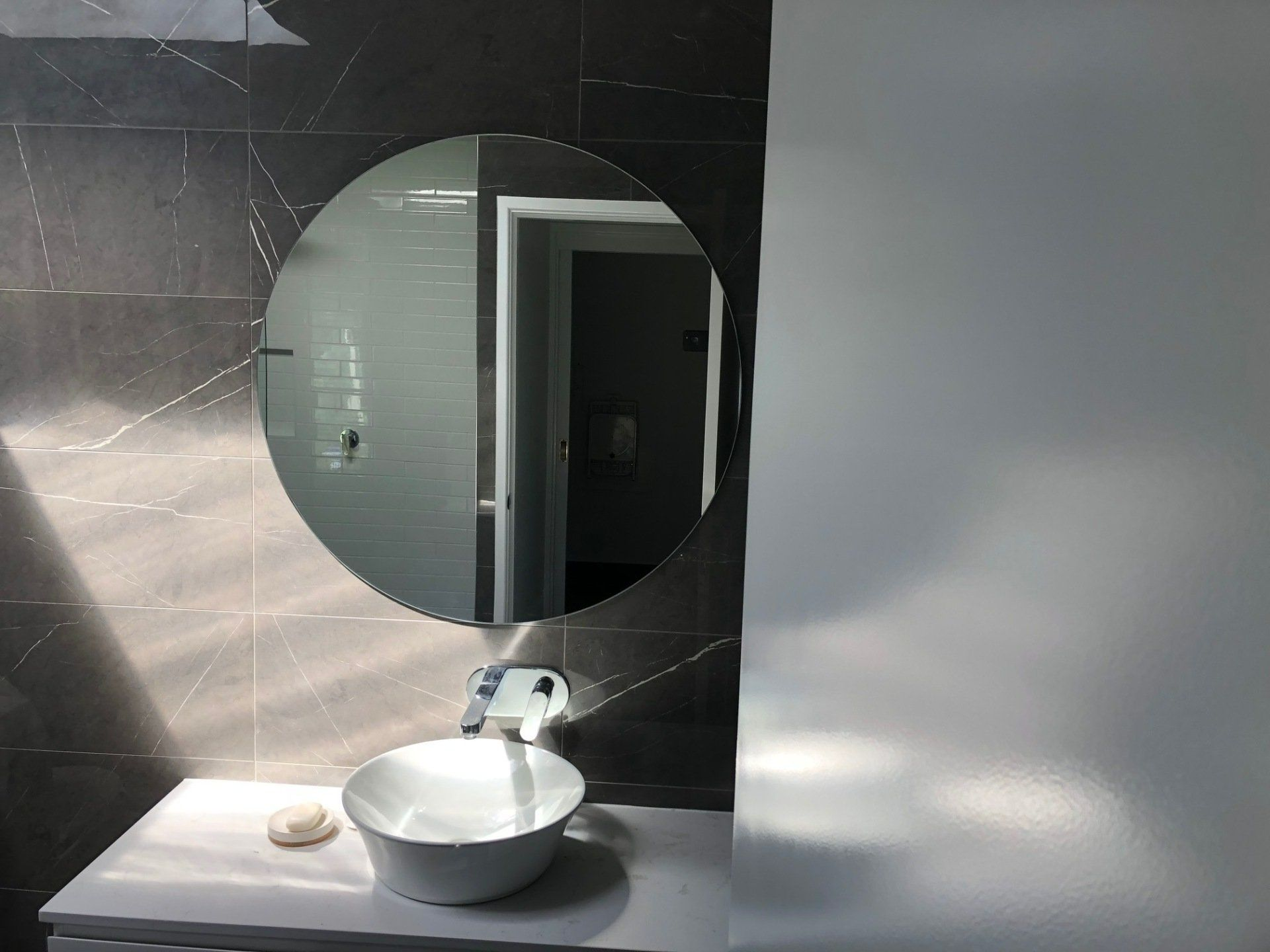Modern bathroom vanity with round mirror, white sink, and dark tiled wall behind it