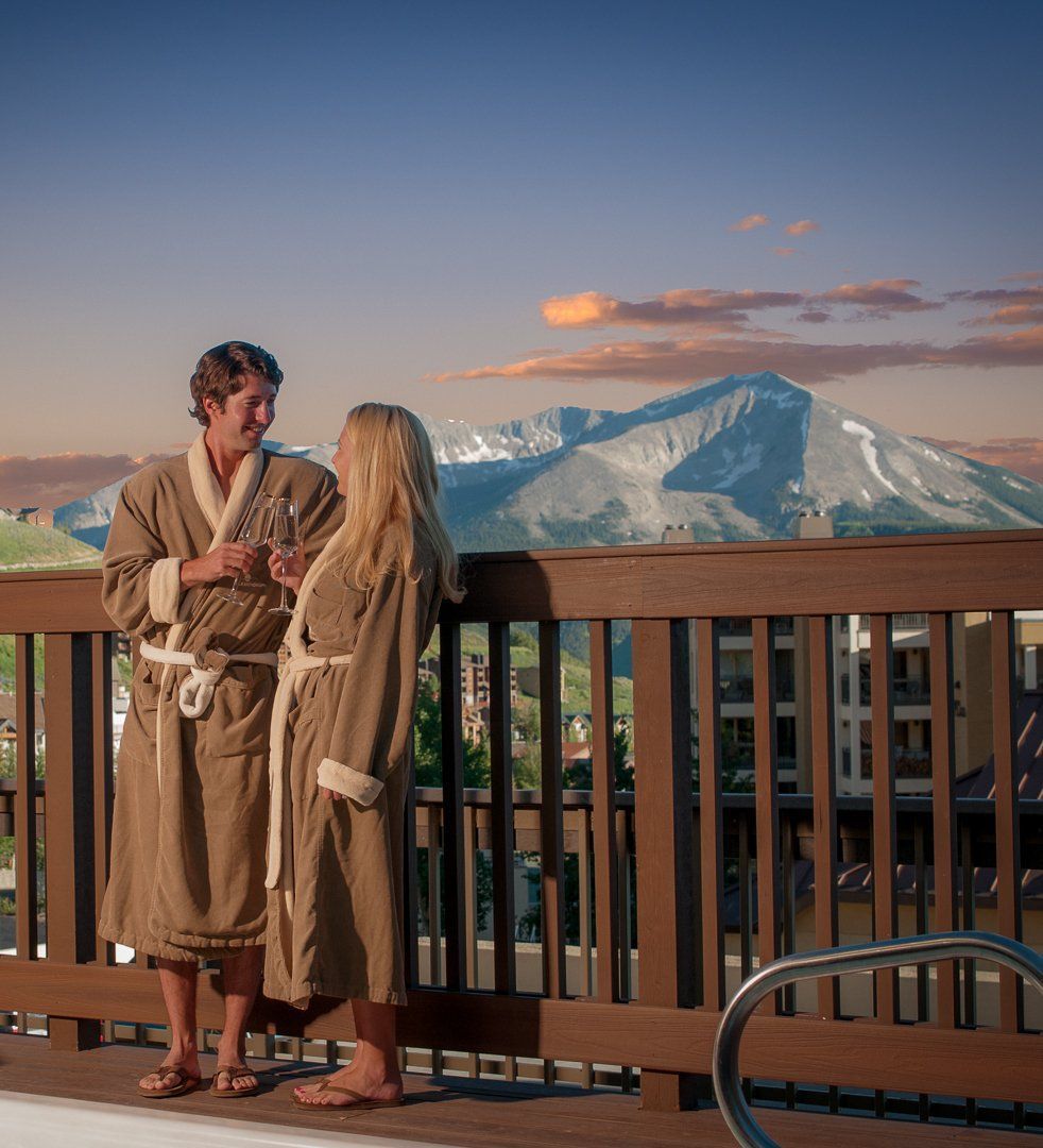 Couple in robes on a deck with mountain view, holding drinks, enjoying the sunset.