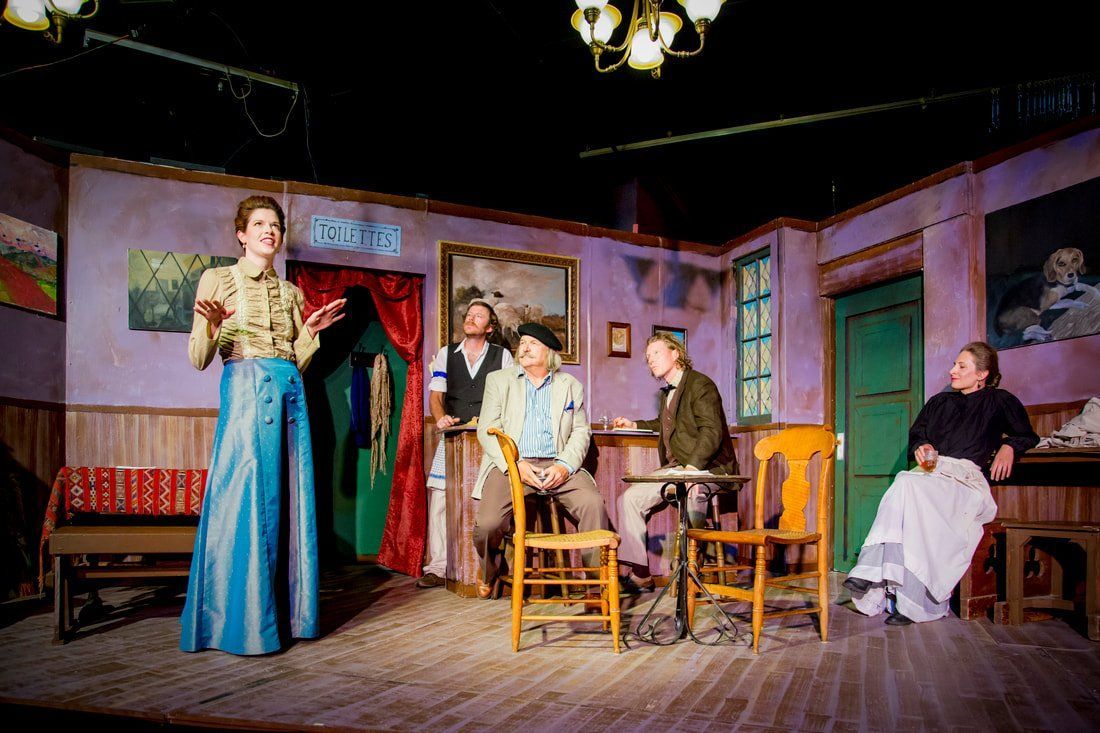 A stage play with six actors, set in a room with period decor and costumes; one woman stands speaking.