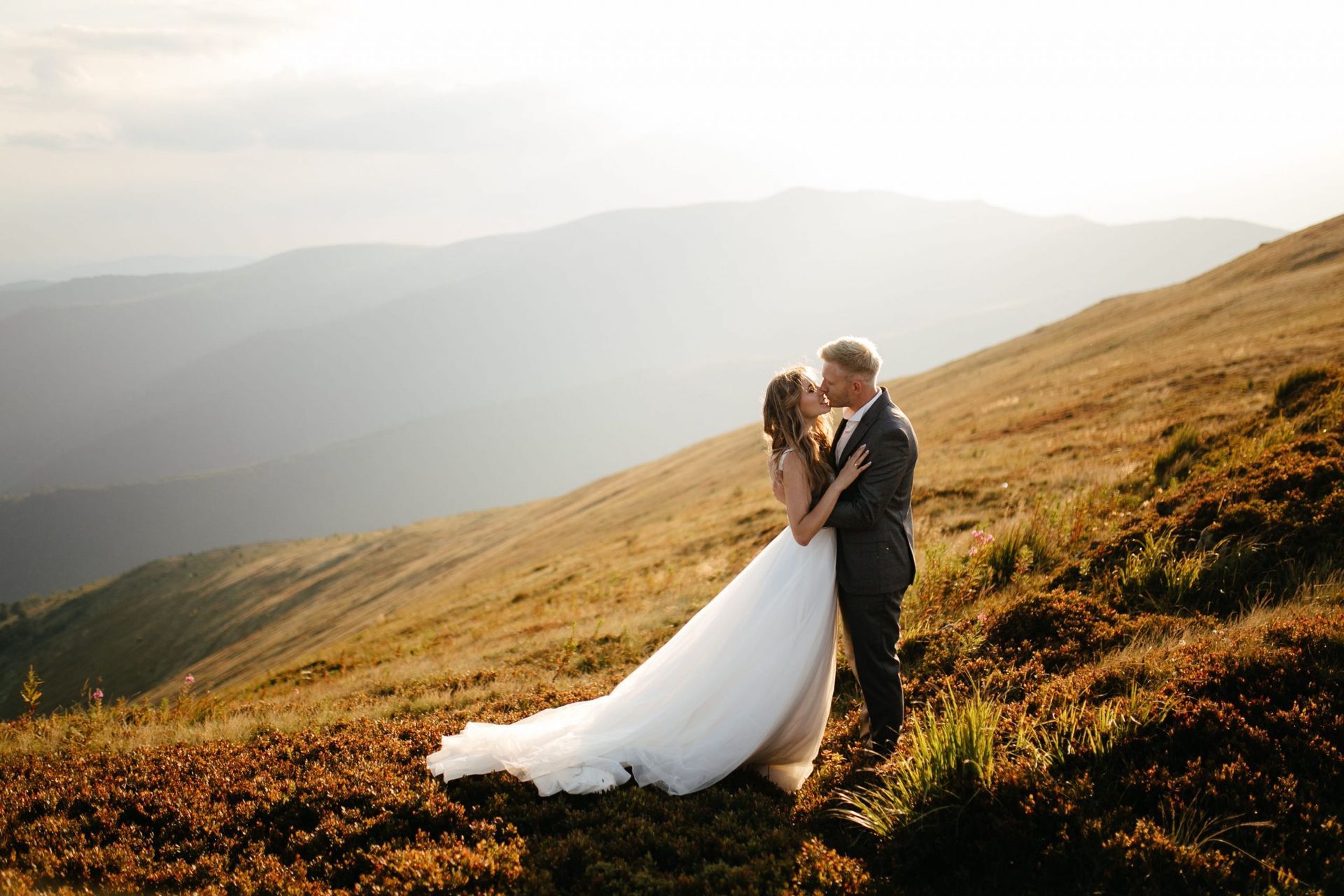 Bride and groom embrace and kiss on a mountain, overlooking a scenic landscape at sunset.