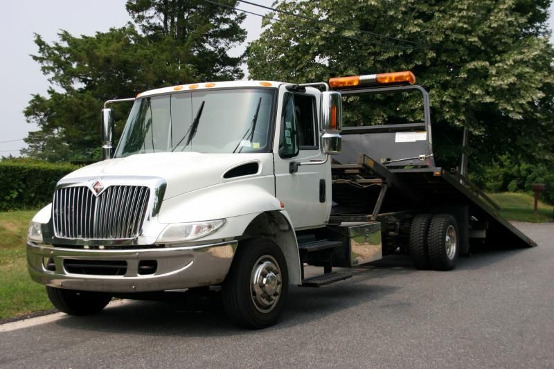 White tow truck on a paved road, parked with the ramp lowered. Orange light bar on cab.