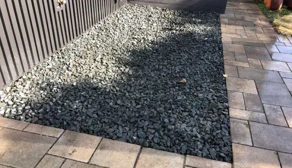 Gravel bed next to a paved walkway and a slatted fence. The gravel is dark, the pavement is light.