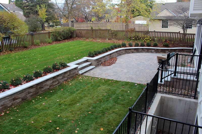 Backyard with green grass, stone patio, and retaining wall with shrubs. Wooden fence in background.