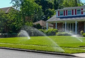 Sprinklers watering a lush green lawn in front of a house with red shutters on a sunny day.
