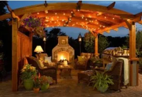 Outdoor seating area under a pergola lit with string lights. Features a fireplace, chairs, and plants.