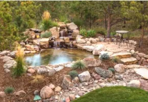A small pond and waterfall feature surrounded by rocks, plants, and a small patio area.