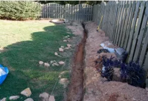 A trench dug along a fence in a backyard, beside a lawn with rocks.