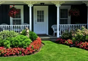 Green lawn and colorful flowerbed lead to a white-doored home with a porch, hanging flowers.