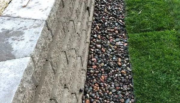 Gray stone wall with a bed of dark pebbles next to green grass.