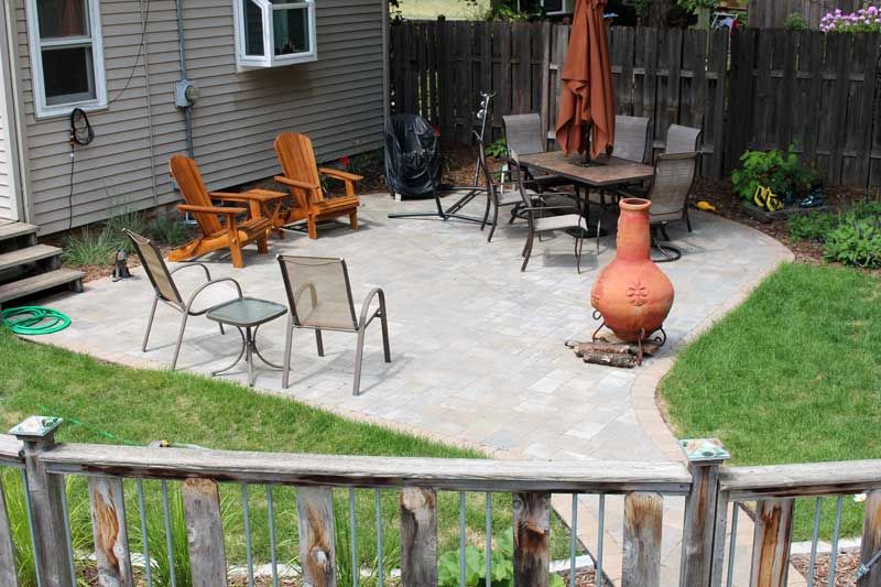 Patio with outdoor furniture: chairs, table, and umbrella. Lawn surrounds a brick patio.