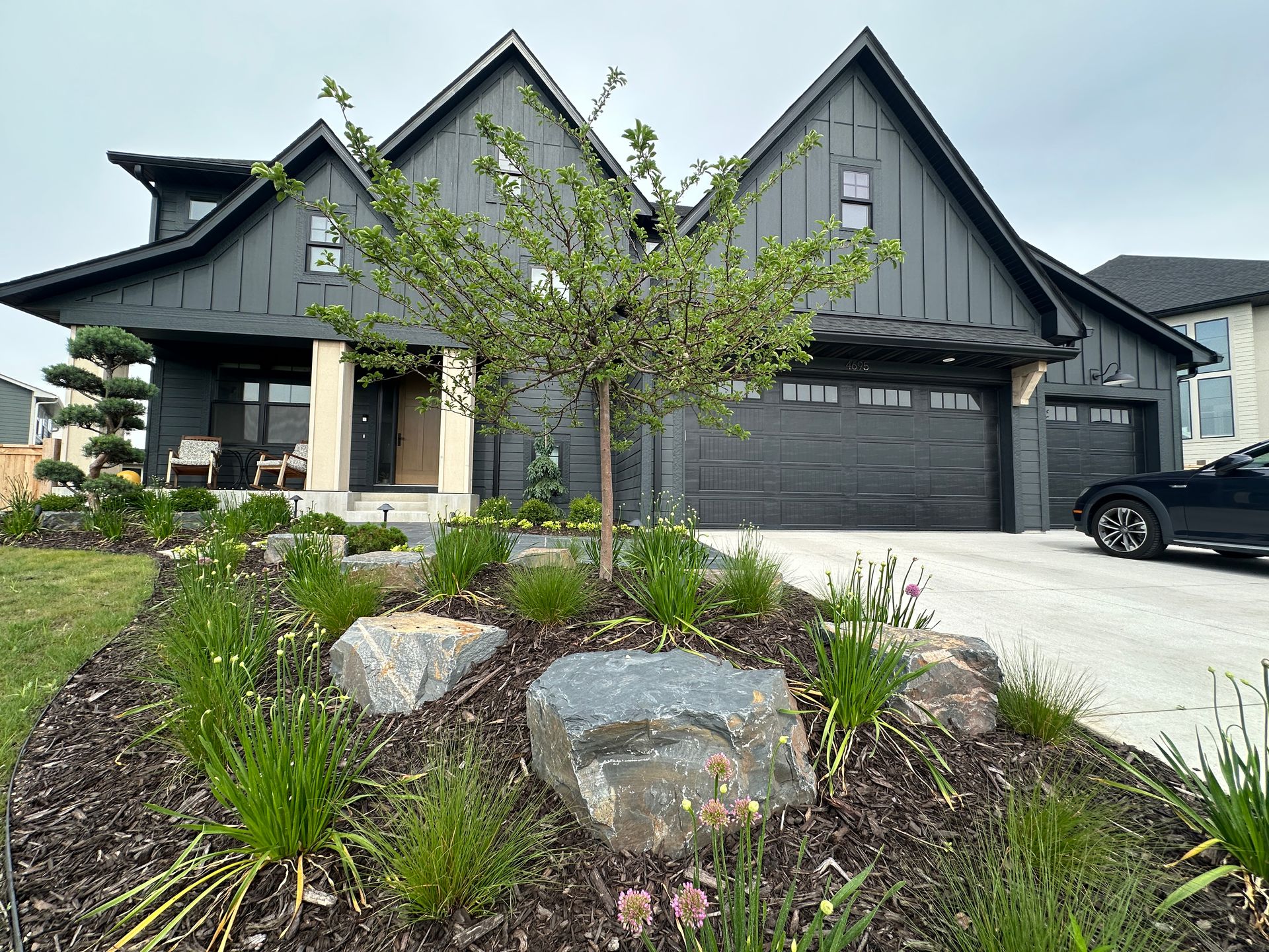 A large house with a lush green lawn and flowers in front of it. Front yard landscaping boulders, Tina crab, allium and mulch 