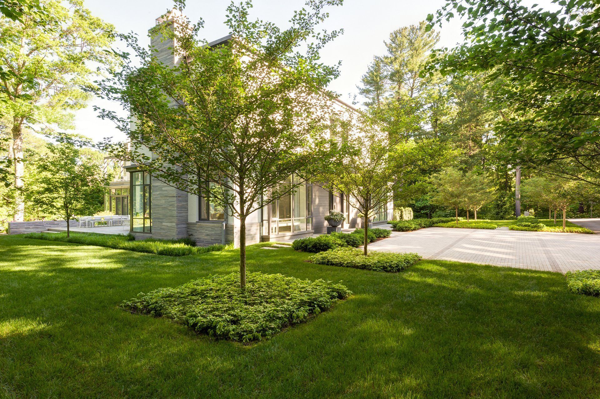 Lush green lawn with trees, house with stone facade, and paved driveway. Sunny, outdoor setting.