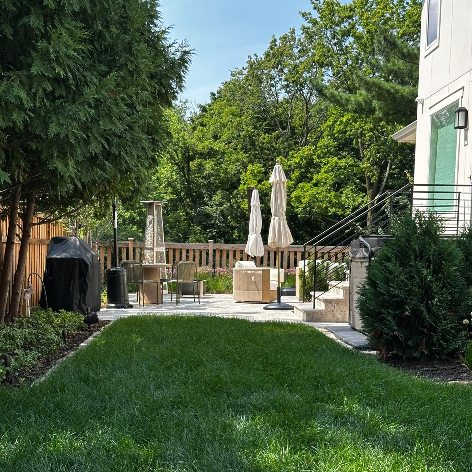 A sunny backyard with a green lawn, a stone patio featuring outdoor seating and umbrellas, and surrounding trees.