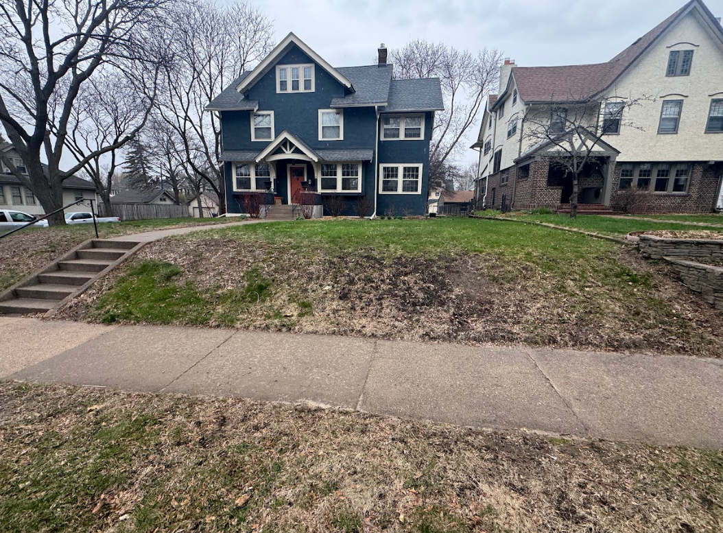 A blue two-story house with a front porch and steps, situated behind a sidewalk and a sloped grassy lawn.