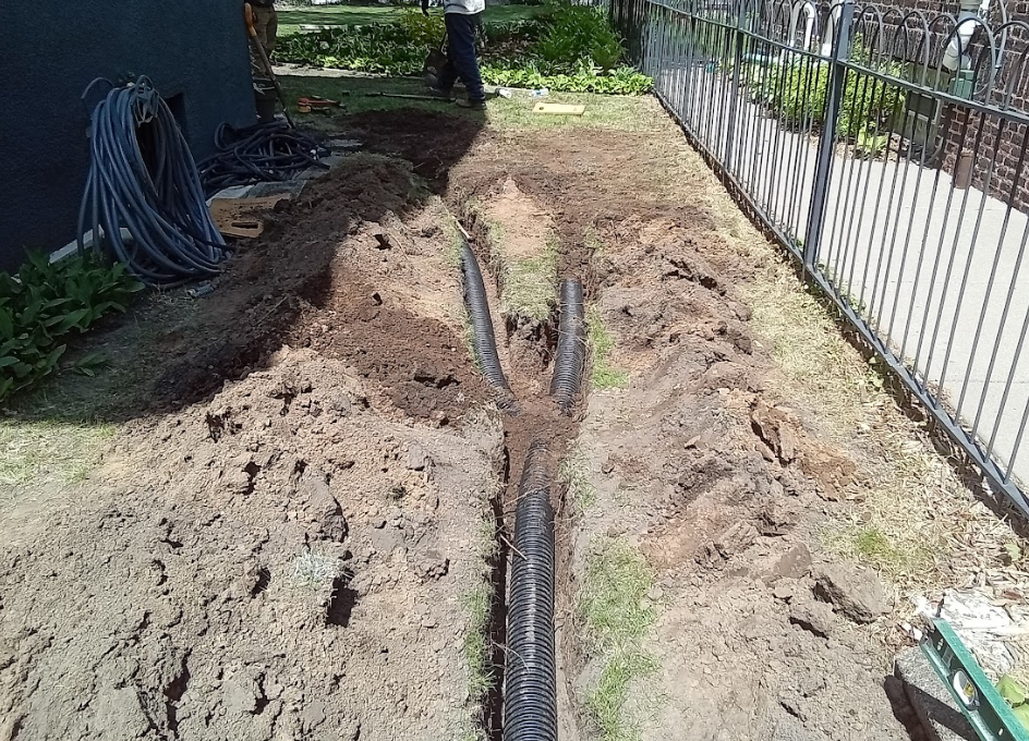 A drainage pipe Y-junction is installed in a trench dug in a residential yard next to a metal fence.
