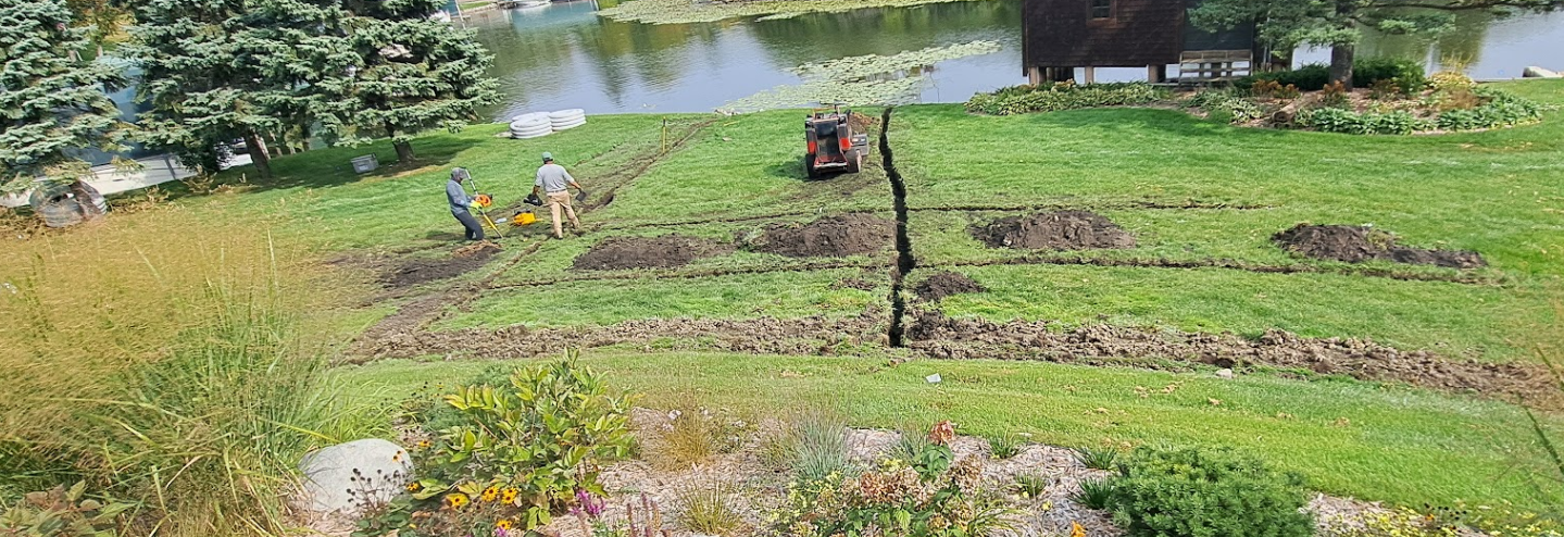 Two people till rows of soil in a grassy yard next to a body of water.