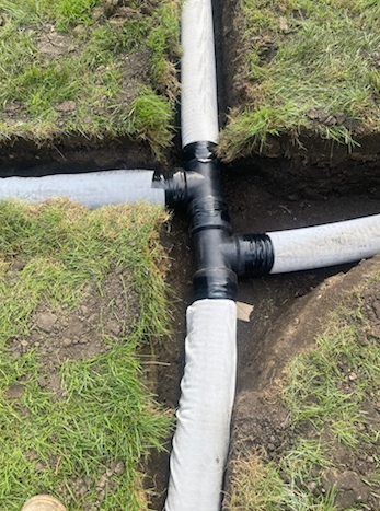 A black plastic T-shaped pipe connector joining three white flexible drainage pipes in a trench cut into a grassy lawn.