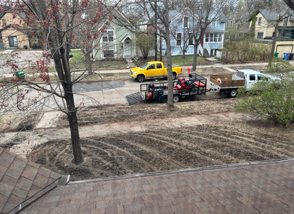 A yellow truck and a work trailer parked on a suburban street next to a front yard with freshly tilled garden beds.
